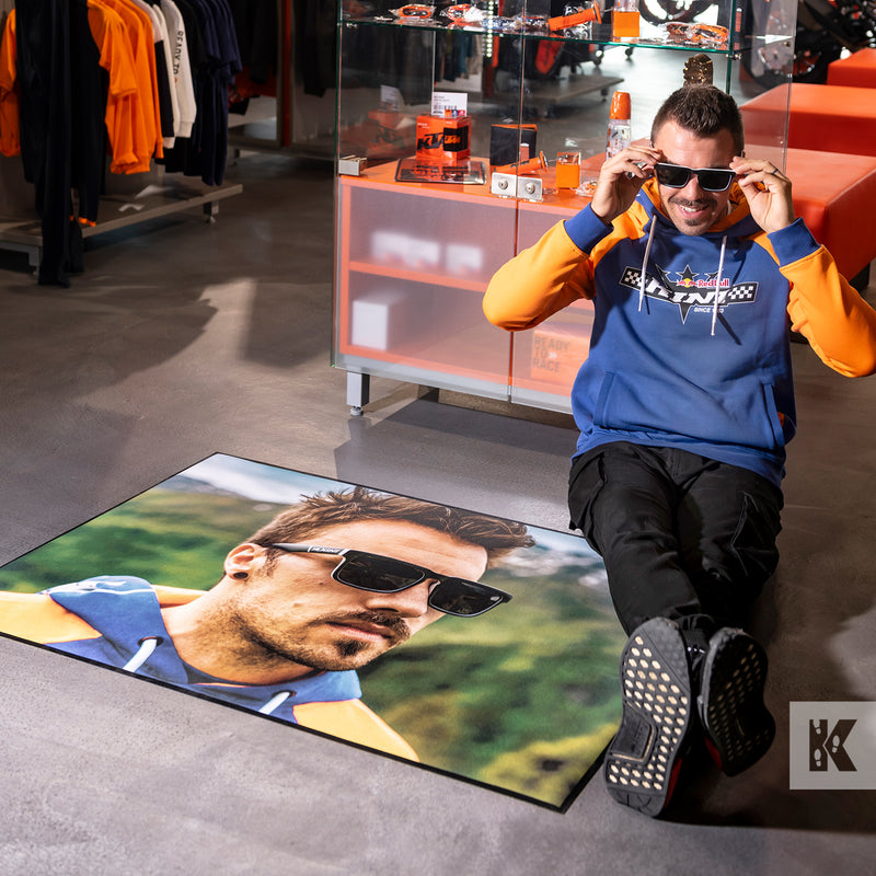 Ad-Mat - Man wearing a RedBull top sitting on the floor while trying on new glasses. The floor has a large Ad-Mat with an image of a man in sunglasses.