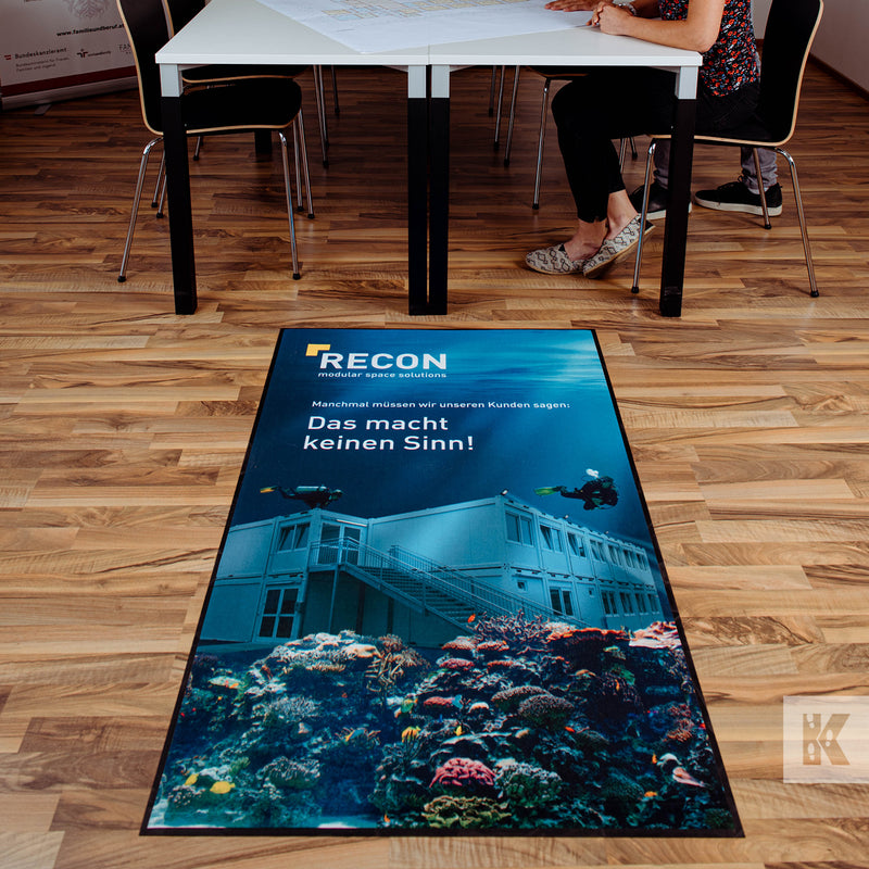 Ad-Mat - Looking down onto an office floor with a white top desk in the background. A mat in the foreground with Recon branding and an underwater scene.