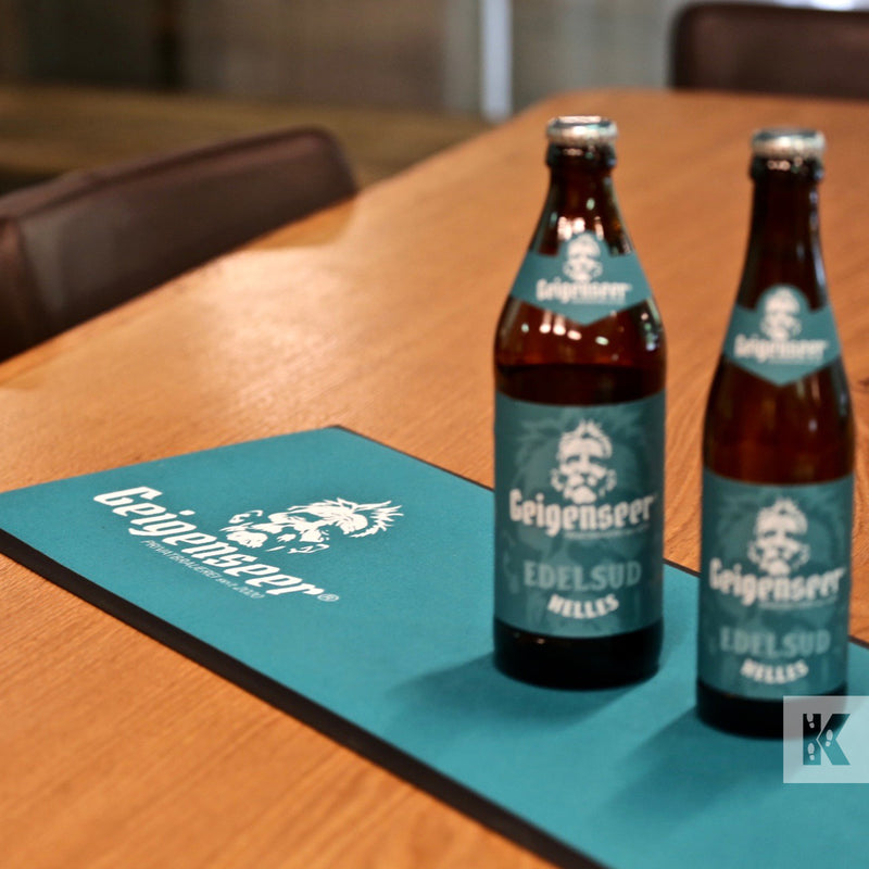 Bar-Mat - Two bottles of Geigenseer beer stood on a mat with the same branding graphics on a polished wooden table top