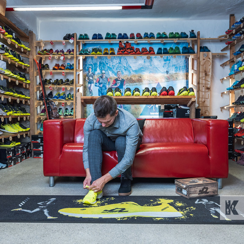 Jet-Print Velour - A man trying on a new pair of trainers, sitting on a red leather couch in a shoe store with a printed in-store promotional mat on the floor