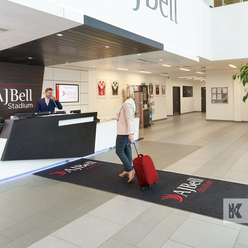 Jet-Print Horizon - Woman arriving at AJ Bell reception, pulling a small red suitcase across a branded runner mat