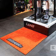 An orange shaggy doormat with a black and white BIKEQUADRAT logo lies on a dark gray tiled floor in front of a Lenz indoor cycling platform, with a person's legs and feet visible stepping onto the platform.