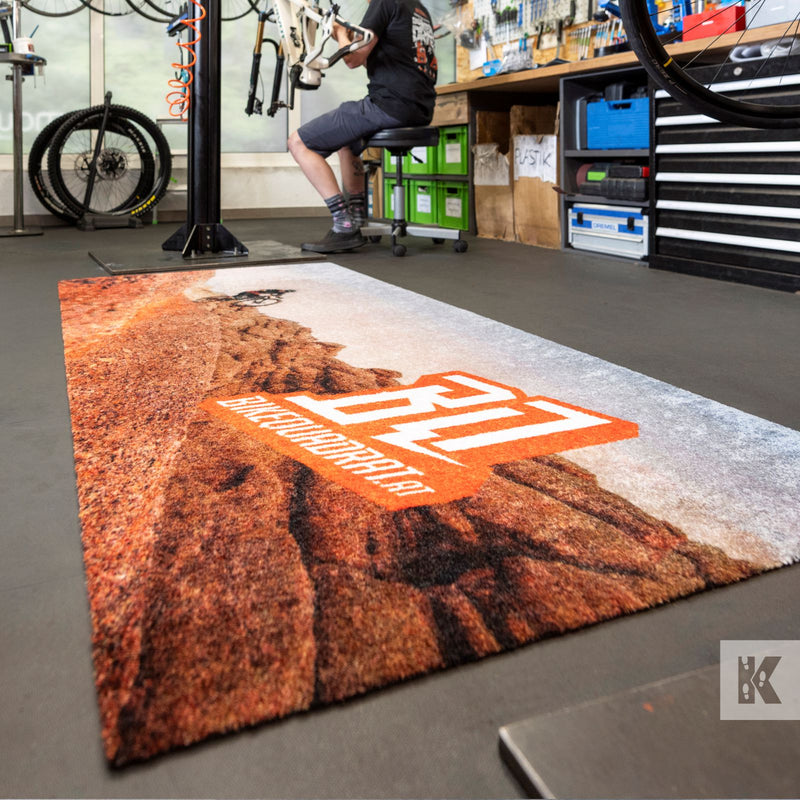 A rectangular shag rug with a desert canyon landscape print and a large orange 'BQ' logo, shown on a workshop floor with a person sitting on a bike stand in the background.