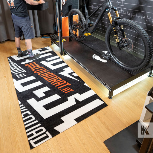 A black and white patterned runner rug with orange text reading 'BIKEQUADRAT' is shown on a wooden floor in a bike shop, next to a mountain bike on a trainer. A person is partially visible on the left.