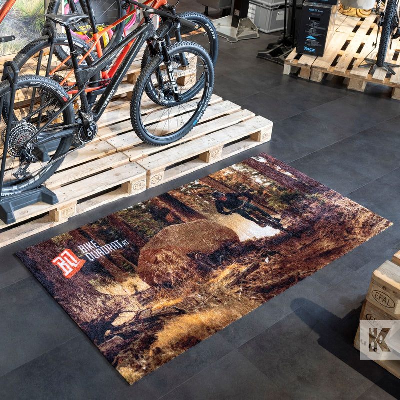 A rectangular area rug with a rustic mountain biking forest scene print, displayed on a dark gray tiled floor in a bike shop next to wooden display stands with bicycles.