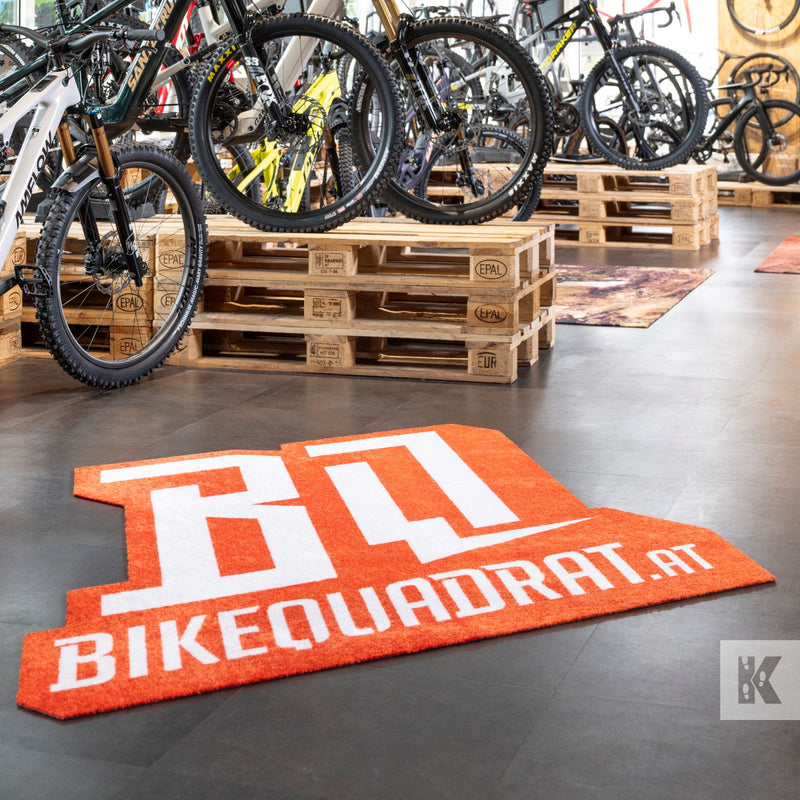 An orange and white textured floor mat with the text 'BIKEQUADRAT.AT' in large white letters, placed on a dark floor inside a bike shop with bicycles and wooden pallets in the background.