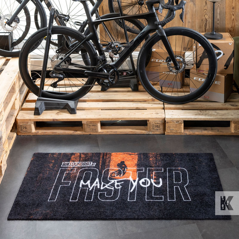 A dark gray and orange textured doormat with 'MAKE YOU FASTER' text and a cyclist silhouette, displayed on a gray floor in front of wooden pallets and black road bicycles.