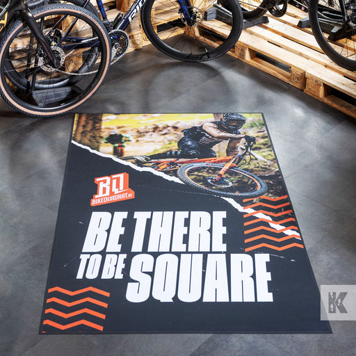 A rectangular printed floor mat or rug displayed on a gray floor in a bike shop, featuring a mountain biker photo, orange and white zigzag graphics, and the text 'BE THERE TO BE SQUARE' with a visible 'BQ' brand logo.