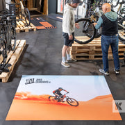 A large rectangular floor mat with a mountain bike rider graphic and the text 'BIKE QUADRAT' is displayed on a dark tiled showroom floor, with two men standing nearby looking at bicycles.