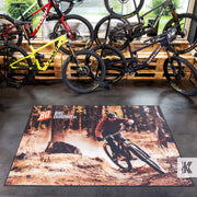 A large rectangular floor mat featuring a mountain biking scene in a forest, displayed on a showroom floor in front of several mountain bikes.