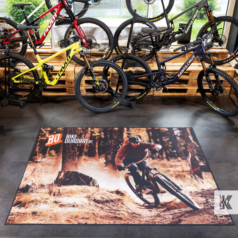 A large rectangular floor mat featuring a mountain biking scene in a forest, displayed on a showroom floor in front of several mountain bikes.