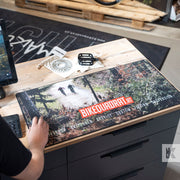 A large rectangular desk mat featuring a forest trail graphic and the text 'BIKEQUADRAT.AT' is laid on a wooden workbench.