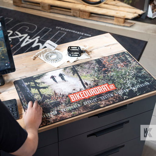 A large rectangular desk mat featuring a forest trail graphic and the text 'BIKEQUADRAT.AT' is laid on a wooden workbench.