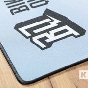 A close-up of a light blue gaming mousepad with a black and white stylized logo, resting on a light wood surface.