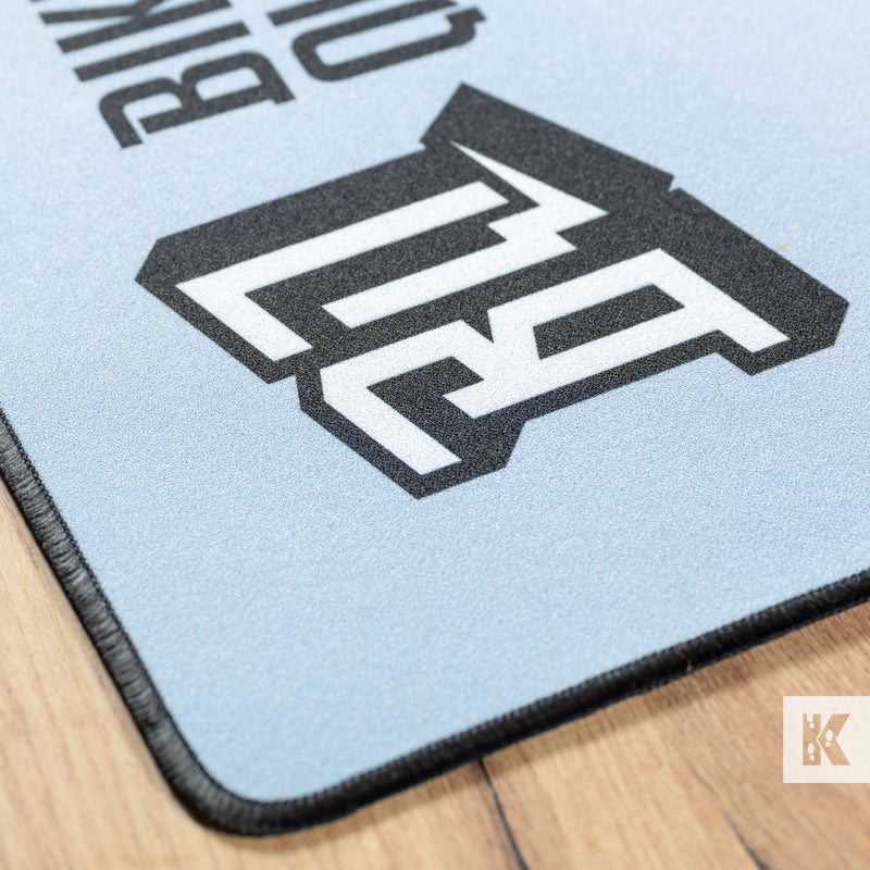 A close-up of a light blue gaming mousepad with a black and white stylized logo, resting on a light wood surface.