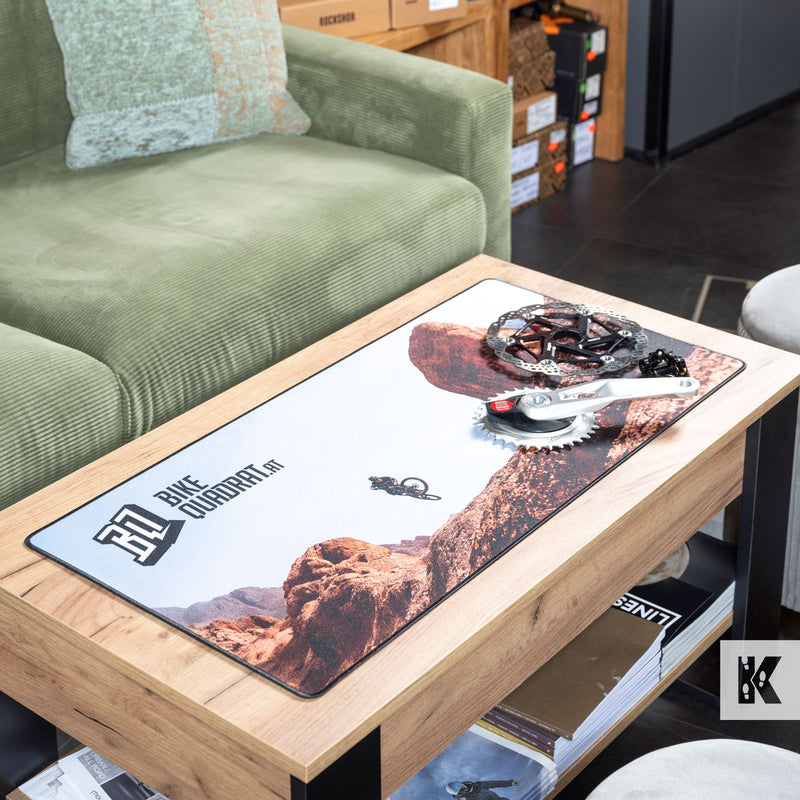 A rectangular desk mat with a desert canyon landscape print and the text 'Bike Outdoors' lies on a wooden coffee table. A bicycle crankset rests on top of the mat, and the setting includes a green sofa and a shelf in the background.