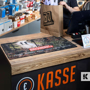 A close-up of a retail counter with a large printed mousepad featuring a forest path and the text 'BIKEQUADRAT'. A person in a black shirt holds a brown paper bag with the Bikequadrat logo behind the counter.