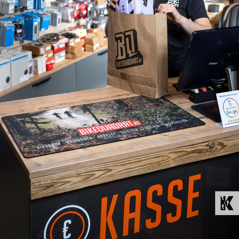 A close-up of a retail counter with a large printed mousepad featuring a forest path and the text 'BIKEQUADRAT'. A person in a black shirt holds a brown paper bag with the Bikequadrat logo behind the counter.